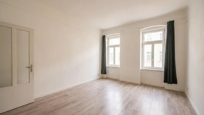 Helles Zimmer mit hellem Laminatboden, weißen Wänden und zwei hohen Fenstern.