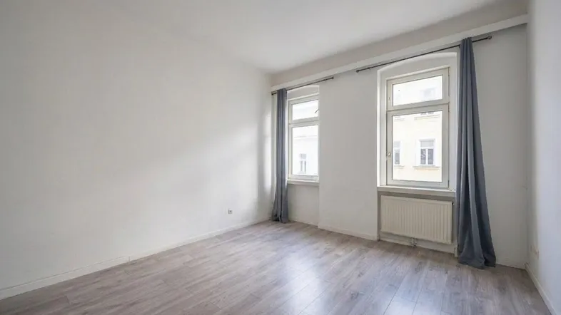 Helles Zimmer mit modernem Laminatboden, weißen Wänden und zwei Fenstern mit grauen Vorhängen.