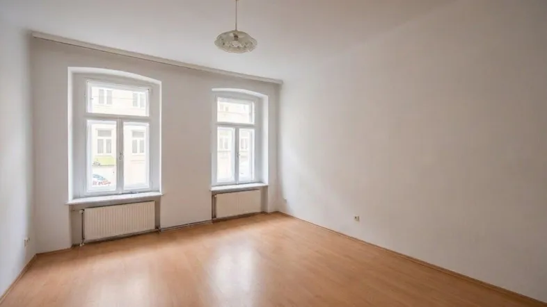 Helles, leeres Zimmer mit zwei Fenstern, Parkettboden und Heizkörpern.