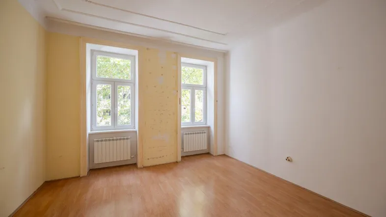 Helles Zimmer mit zwei großen Fenstern, Holzboden und hohen Decken, bereit zur Renovierung.
