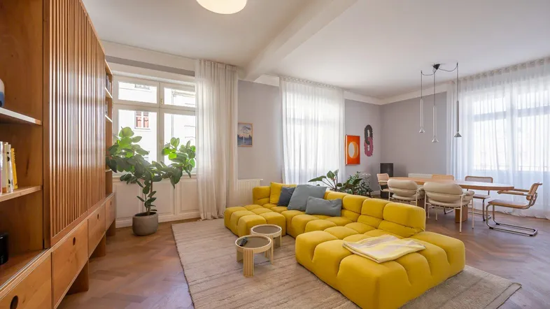 Helles Wohnzimmer mit gelbem Sofa, Holzbücherregal und Parkettboden, offen zum Essbereich.