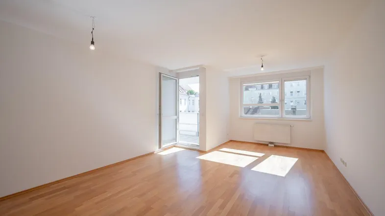 Helles, leeres Zimmer mit Parkettboden und Zugang zu einem Balkon, gut beleuchtet.