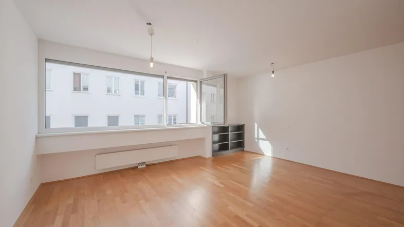 Helles, leeres Wohnzimmer mit Parkettboden und großen Fenstern, die viel Tageslicht hereinlassen.