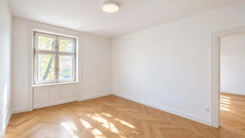 Heller, leerer Raum mit Fischgrätparkett, weißen Wänden und viel natürlichem Tageslicht.