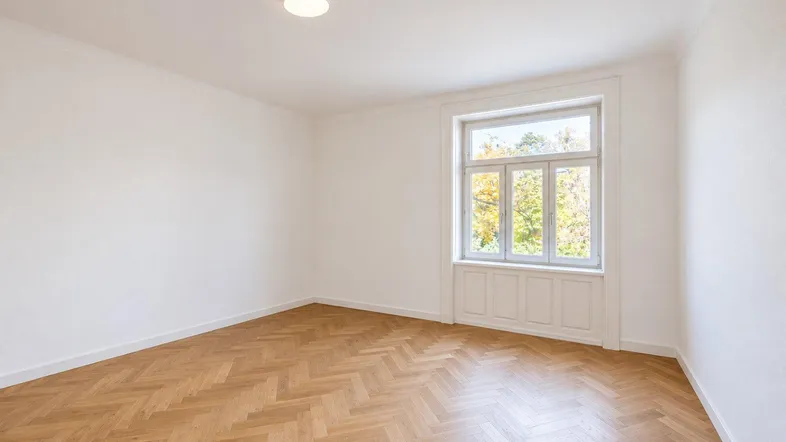 Heller Raum mit klassischem Fischgrätparkett, hohen Decken und Blick ins Grüne durch das Fenster.