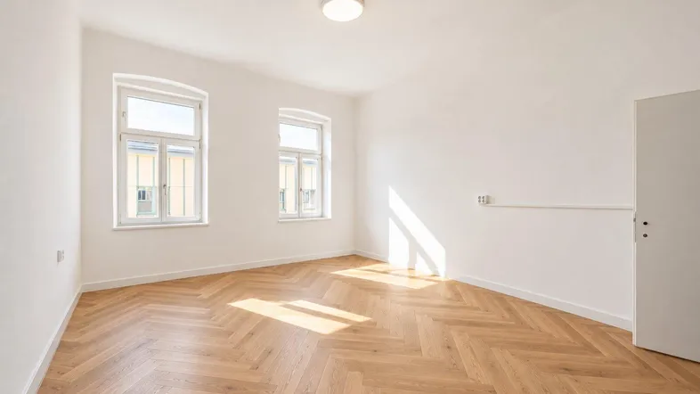 Helles, leeres Zimmer mit Fischgrätparkett und zwei großen Fenstern, das viel Tageslicht hereinlässt.