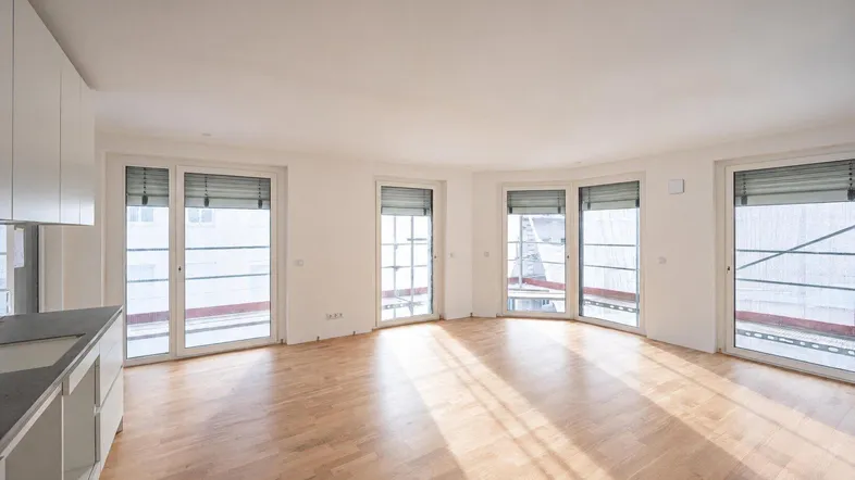Helles, offenes Zimmer mit Holzboden und mehreren Fenstertüren, die viel Tageslicht hereinlassen.