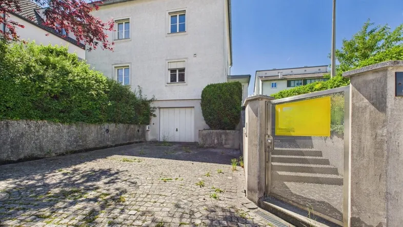 Weißes Mehrfamilienhaus mit integrierter Garage, gepflasterter Einfahrt und grünen Hecken.