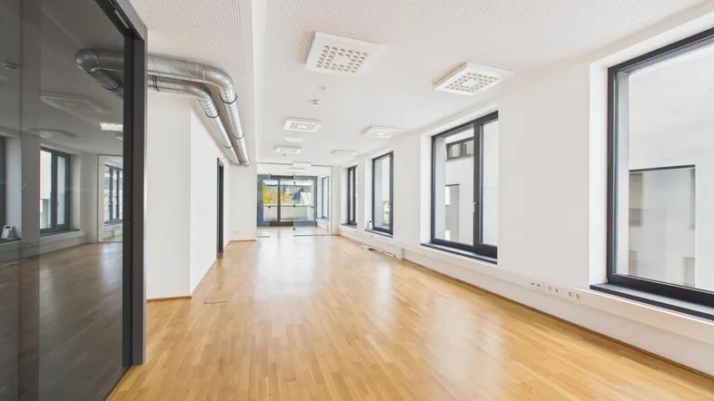Heller, langer Büroraum mit Holzboden, großen Fenstern und sichtbaren Lüftungsrohren an der Decke.