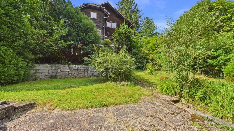 Gartenbereich mit Rasenfläche, Steinmauer und Blick auf das Haus durch die Bäume.