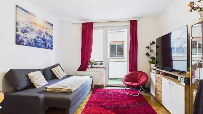 Helles Wohnzimmer mit Sofa, Fernseher, rotem Teppich und Zugang zum Balkon.