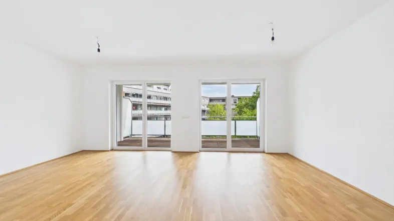 Heller, leerer Wohnraum mit neuem Holzfußboden und Zugang zu zwei Balkonen mit Grünblick.