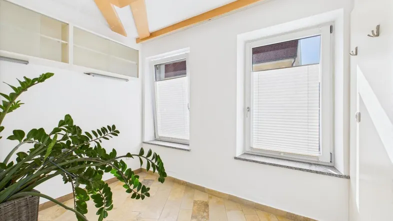 Heller Innenraum mit zwei Fenstern, Jalousien, einem Deckenventilator und hellen Fliesenböden.