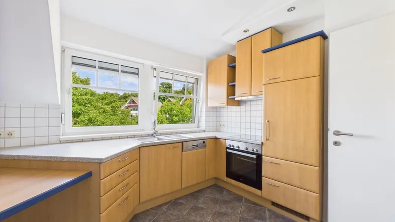 Helle Küche mit Holzschränken, Fliesenboden und einem Fenster mit Blick ins Grüne.