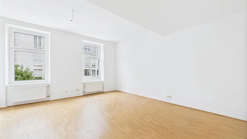 Unmöblierter Raum mit hellen Wänden, Holzfußboden und zwei Fenstern, die Tageslicht hereinlassen.