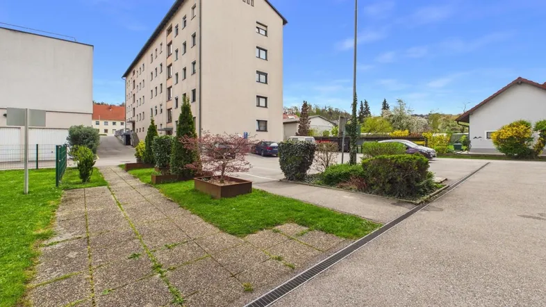 Mehrfamilienhaus mit Parkplätzen, Grünflächen und gepflegter Umgebung unter blauem Himmel.