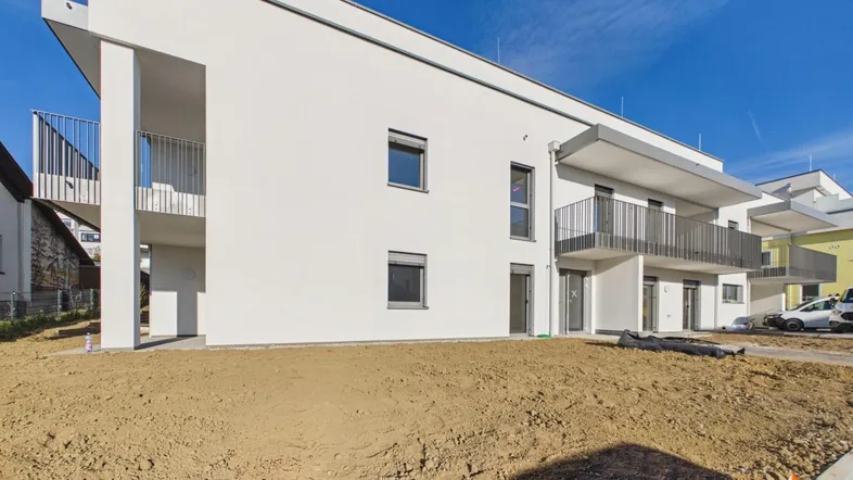 Modernes, weißes Mehrfamilienhaus mit Balkonen und noch unfertiger Außenanlage unter blauem Himmel.