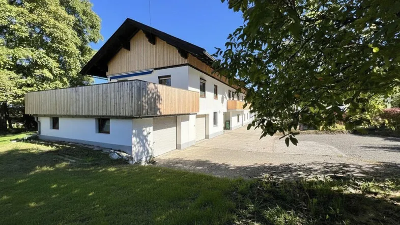 Weißes Gebäude mit Holzverkleidung und Balkonen, umgeben von Grünflächen unter blauem Himmel.