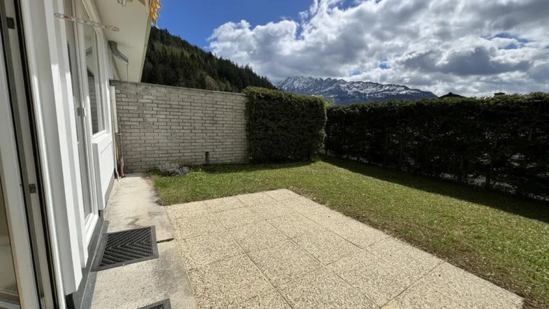 Gepflasterte Terrasse mit Rasenfläche und Blick auf die umliegenden Berge unter bewölktem Himmel.