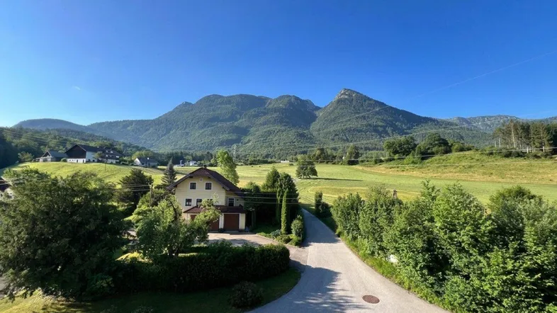 Weitläufige Landschaftsaufnahme mit einem Haus, umgeben von grünen Wiesen und majestätischen Bergen unter blauem Himmel.
