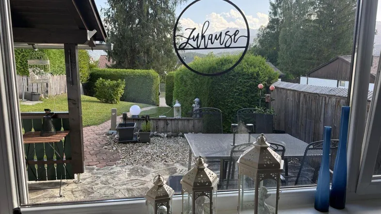 Blick aus dem Fenster auf die Terrasse und den Garten mit Bergpanorama im Hintergrund.