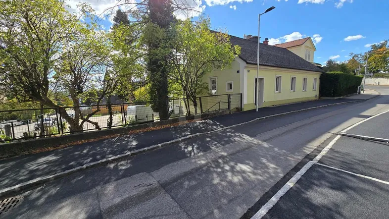 Ansicht des hellgelben Hauses von der Straße mit Bürgersteig und Bäumen unter blauem Himmel.