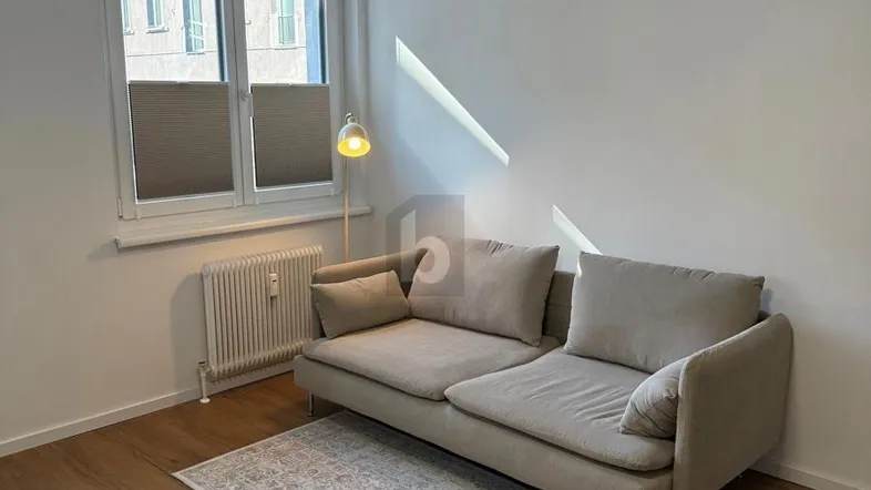 Helles Wohnzimmer mit beigem Sofa, Holzboden und Fenster, das Tageslicht hereinlässt.