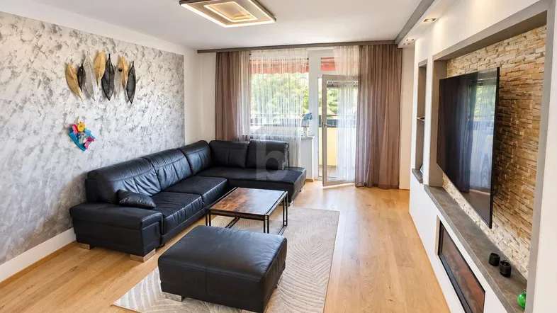 Helles Wohnzimmer mit schwarzem Ecksofa, Holzboden und Zugang zu einem Balkon.
