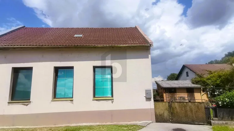 Einfamilienhaus mit beiger Fassade, Satteldach und grüner Umgebung unter bewölktem Himmel.