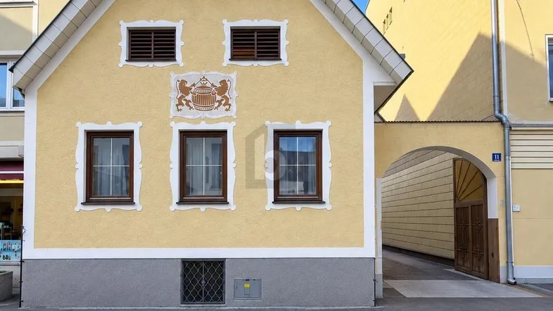 Historisches Gebäude mit gelber Fassade, Satteldach und dekorativem Wappen über dem Eingang.