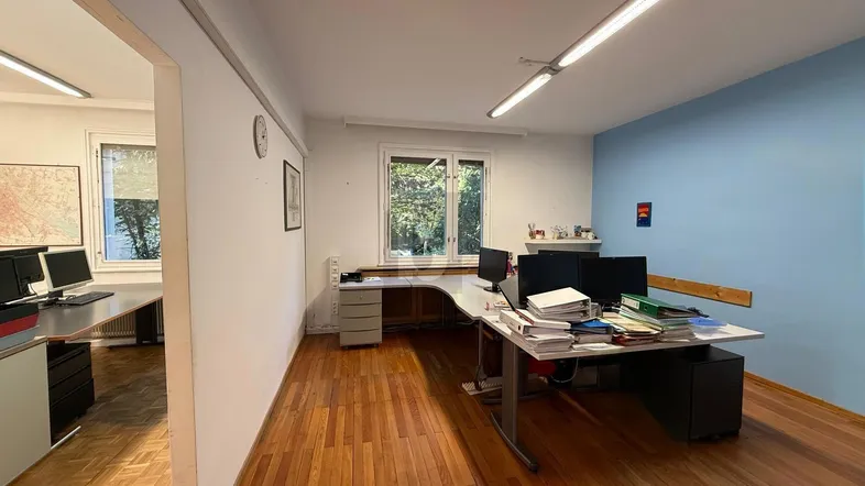 Geräumiges Büro mit Holzparkettboden, einer blauen Akzentwand und viel Tageslicht durch große Fenster.
