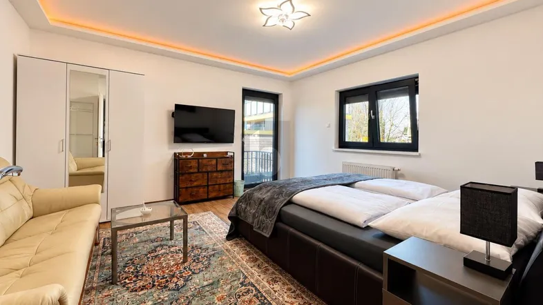 Helles Schlafzimmer mit Doppelbett, Sofa, Fernseher und Zugang zum Balkon.