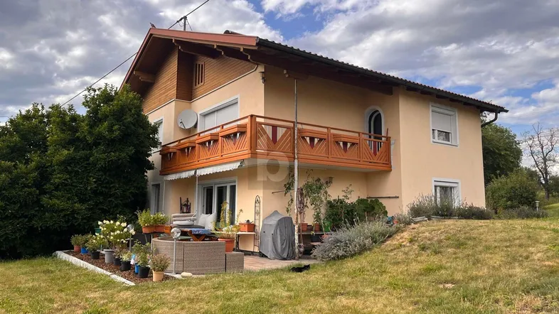 Hausfassade mit Balkon und möblierter Terrasse, umgeben von Grünflächen unter bewölktem Himmel.