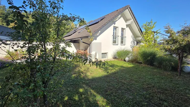 Hausfassade mit gepflegtem Garten, Obstbäumen und Solarpaneelen auf dem Dach unter blauem Himmel.