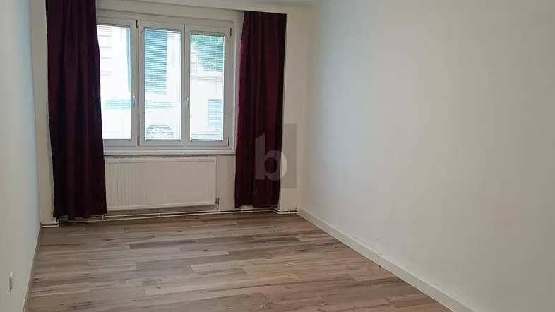 Heller Raum mit Holzoptikboden, weißen Wänden und Fenster mit roten Vorhängen.