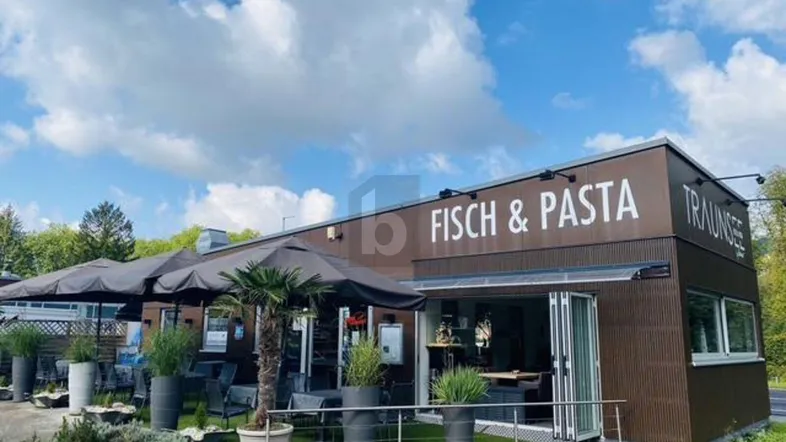 Außenansicht eines Restaurants mit brauner Fassade, großen Sonnenschirmen und einer einladenden Terrasse für Gäste.