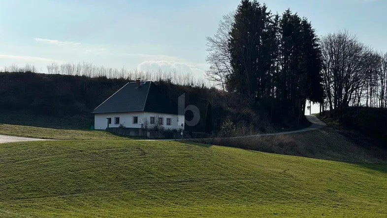 Weißes Haus mit dunklem Dach auf einem grünen Hügel in ländlicher Umgebung.