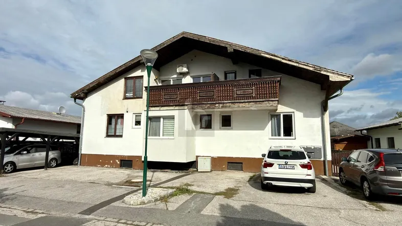 Vorderansicht des Hauses mit Balkon, Satteldach und Stellplätzen im Freien sowie einem Carport.