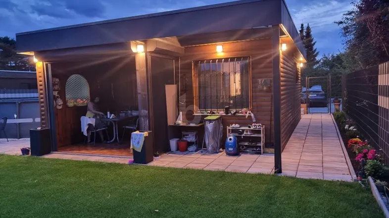 Beleuchtetes Gartenhaus am Abend mit einer kleinen Außenküche, umgeben von Rasen und einem gepflasterten Weg.