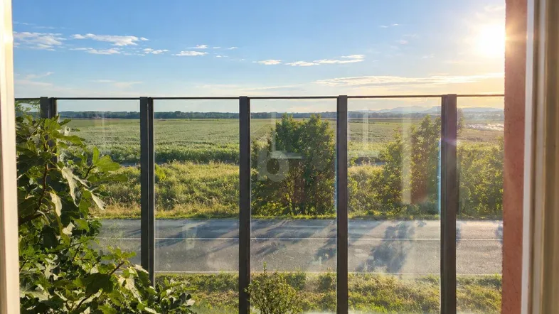 Heller Balkon mit Glasgeländer und weitem Blick über grüne Felder bei Sonnenuntergang.