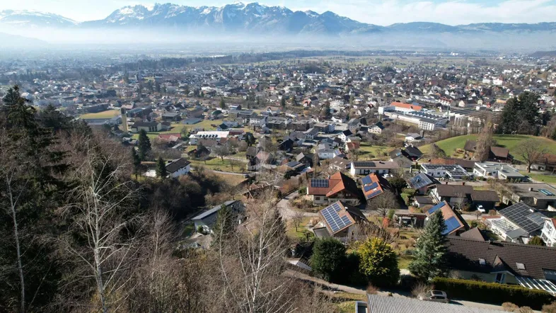 Weitläufige Luftaufnahme einer Siedlung mit grüner Landschaft und majestätischen Bergen im Hintergrund.