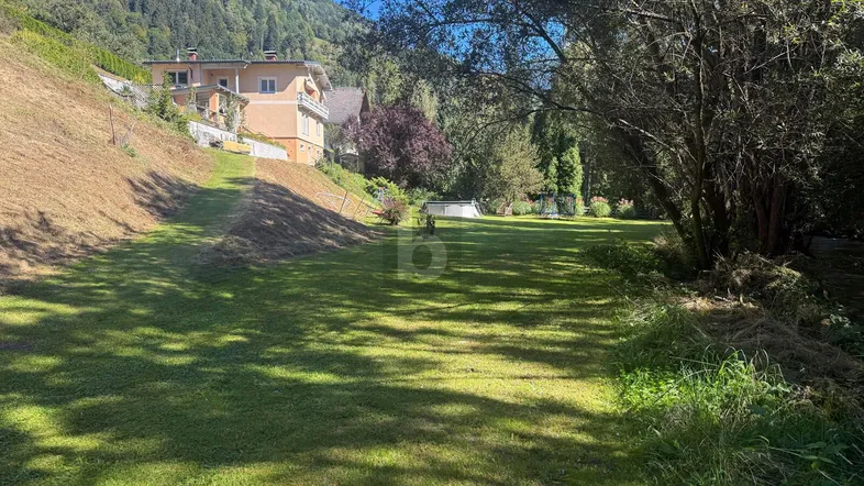 Große Rasenfläche mit einem Haus am Hang und Bergblick, umgeben von Bäumen und Natur.