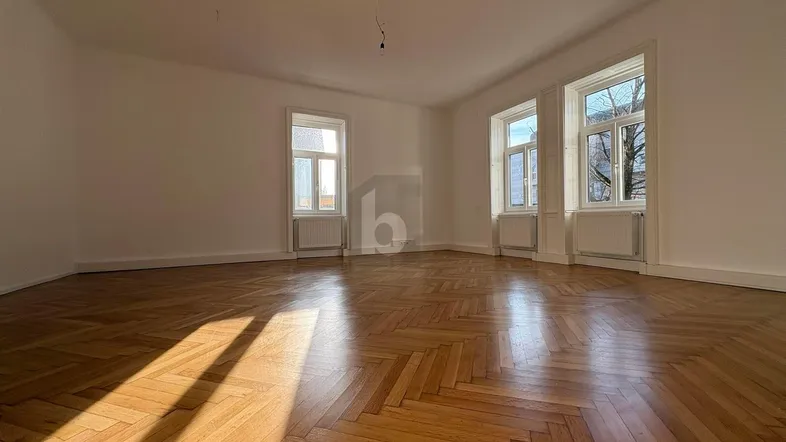 Heller Wohnraum mit klassischem Fischgrätparkett, weißen Wänden und viel Tageslicht durch große Fenster.