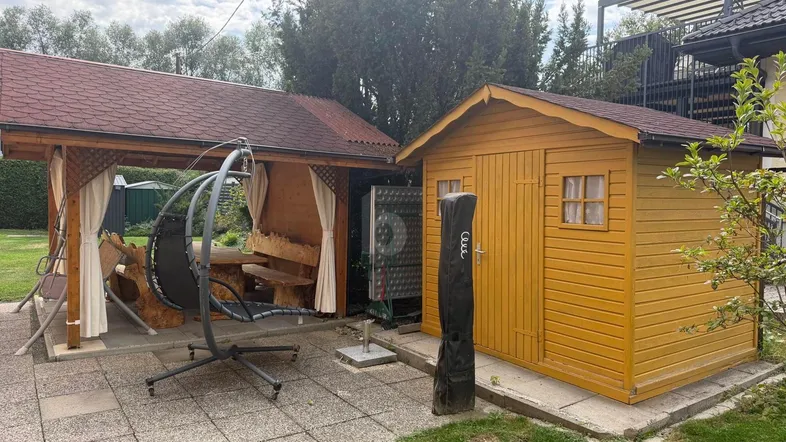 Überdachter Sitzbereich im Garten mit Holzschuppen und Schaukelstuhl, umgeben von Grünflächen.