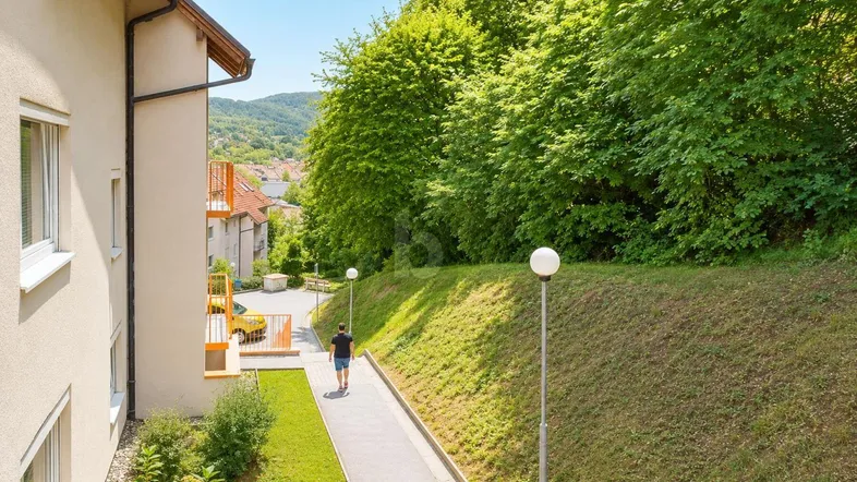 Außenansicht eines Gebäudes mit einem Gehweg und einer grünen, bewaldeten Böschung im Hintergrund.