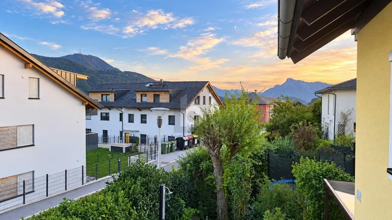 Blick von einem Balkon auf eine Wohnsiedlung mit gepflegtem Garten und majestätischem Bergpanorama bei Sonnenuntergang.