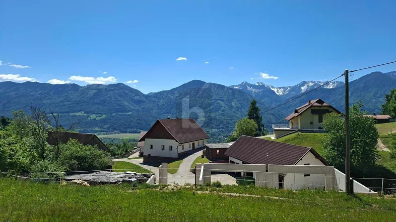 Mehrere Häuser in einer ländlichen Umgebung mit weitem Blick auf schneebedeckte Berge und grüne Hügel unter blauem Himmel.