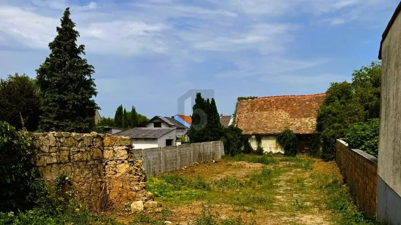 Blick über ein unbebautes Grundstück mit alten Steinmauern und umliegenden Häusern unter blauem Himmel.