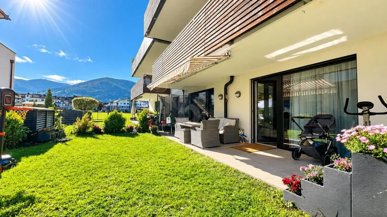 Sonniger Gartenbereich mit gepflegtem Rasen, Terrasse und Bergblick vor moderner Wohnanlage.