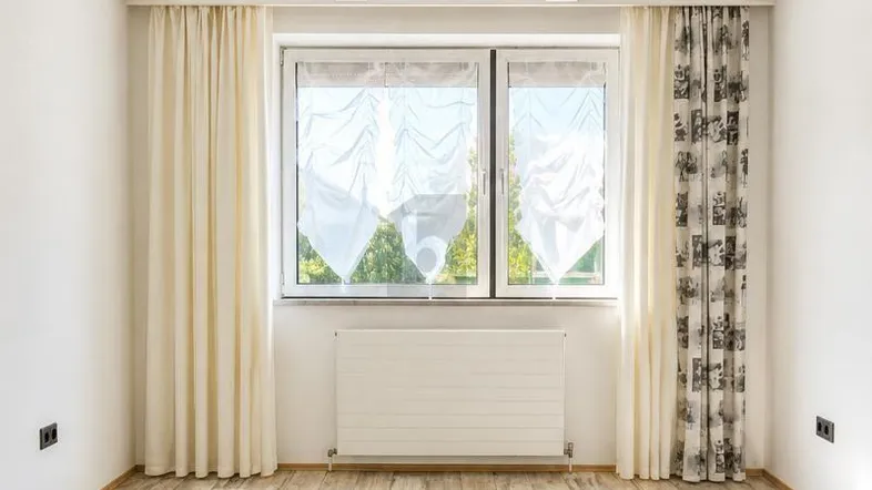 Heller Raum mit Laminatboden, sichtbaren Deckenbalken und großem Fenster mit Heizkörper.